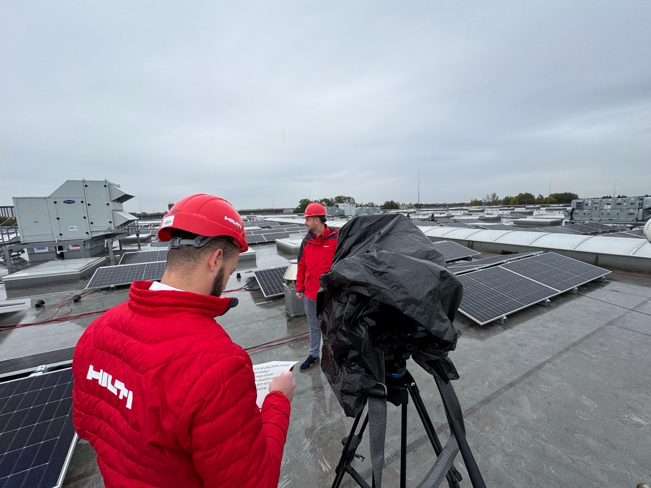 Eine Filmcrew dreht zwischen Solarpaneelen auf einem Dach, Kamera unter Regenschutz.