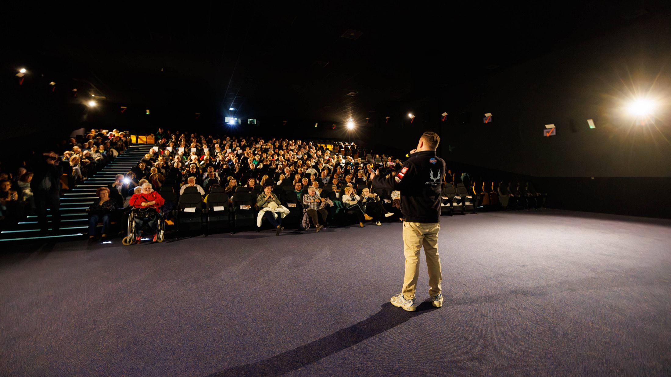 Ein Moderator in ALN-Jacke vor einem vollen, applaudierenden Theaterpublikum unter dramatischen Scheinwerfern.