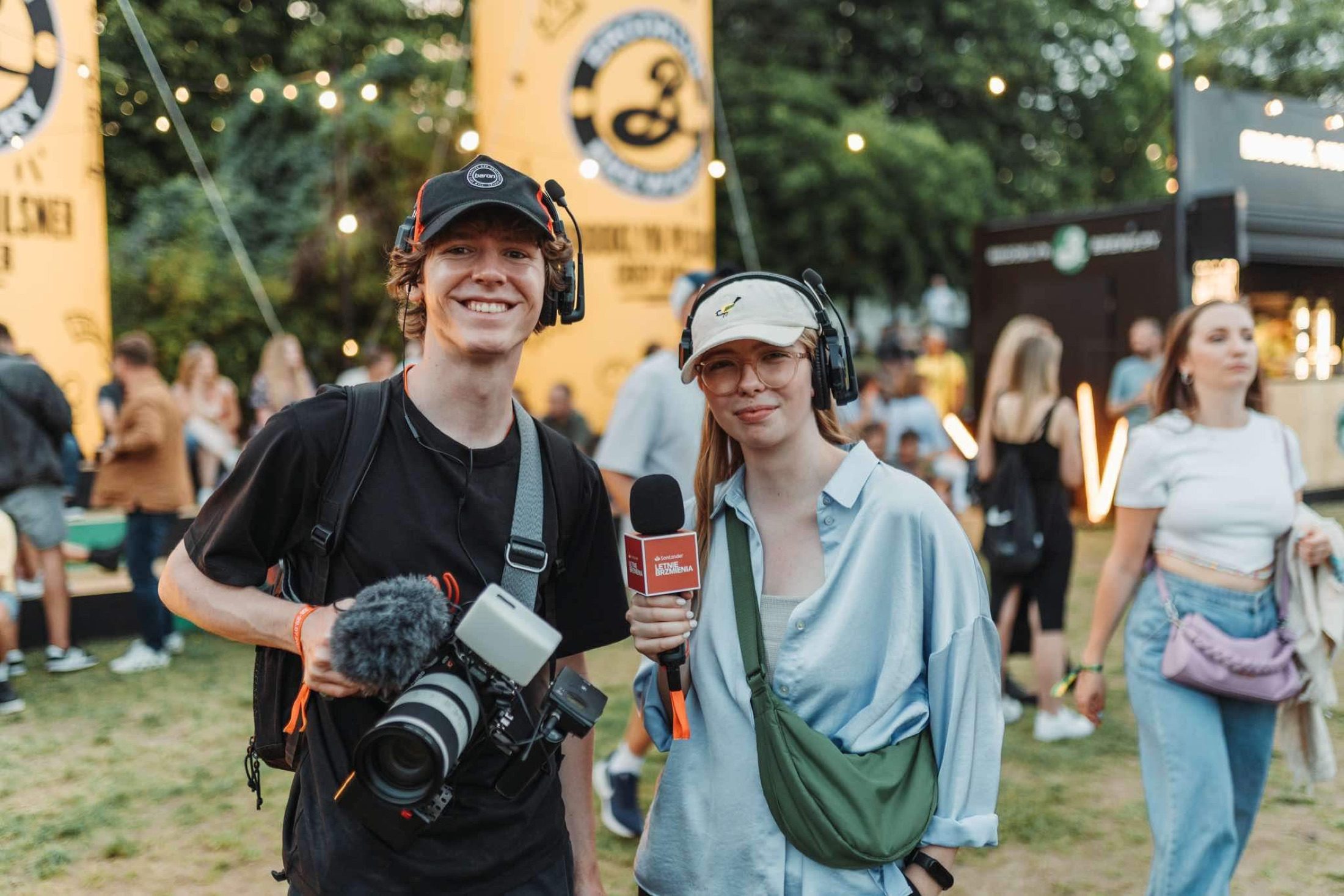 Ein Kameramann und ein Reporter mit gebrandetem Mikrofon, lächelnd auf einem Open-Air-Festival.