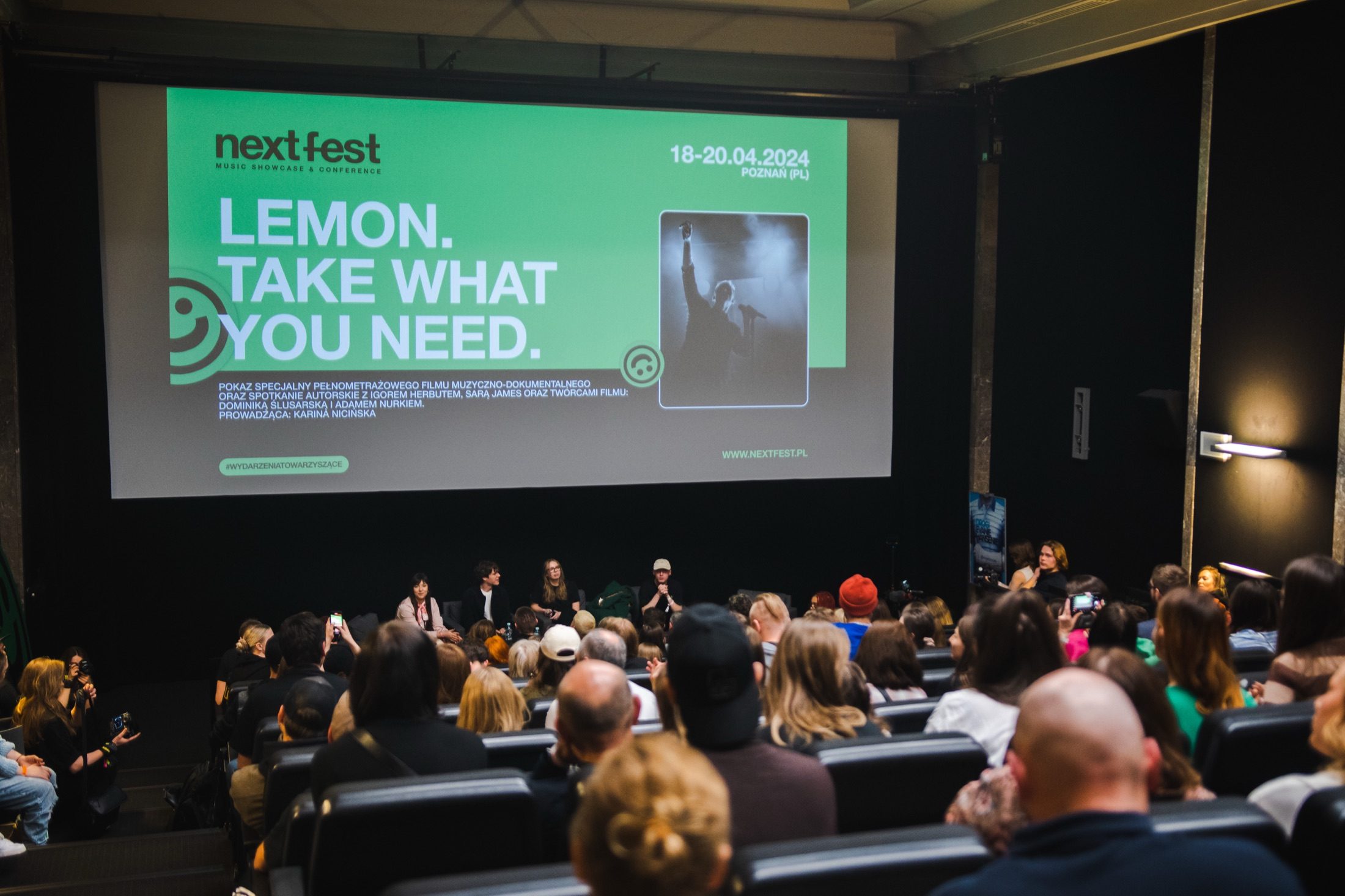 Eine 'LemON. Take what you need.'-Next-Fest-Titeltafel über einem Panel und einem vollen Publikum.