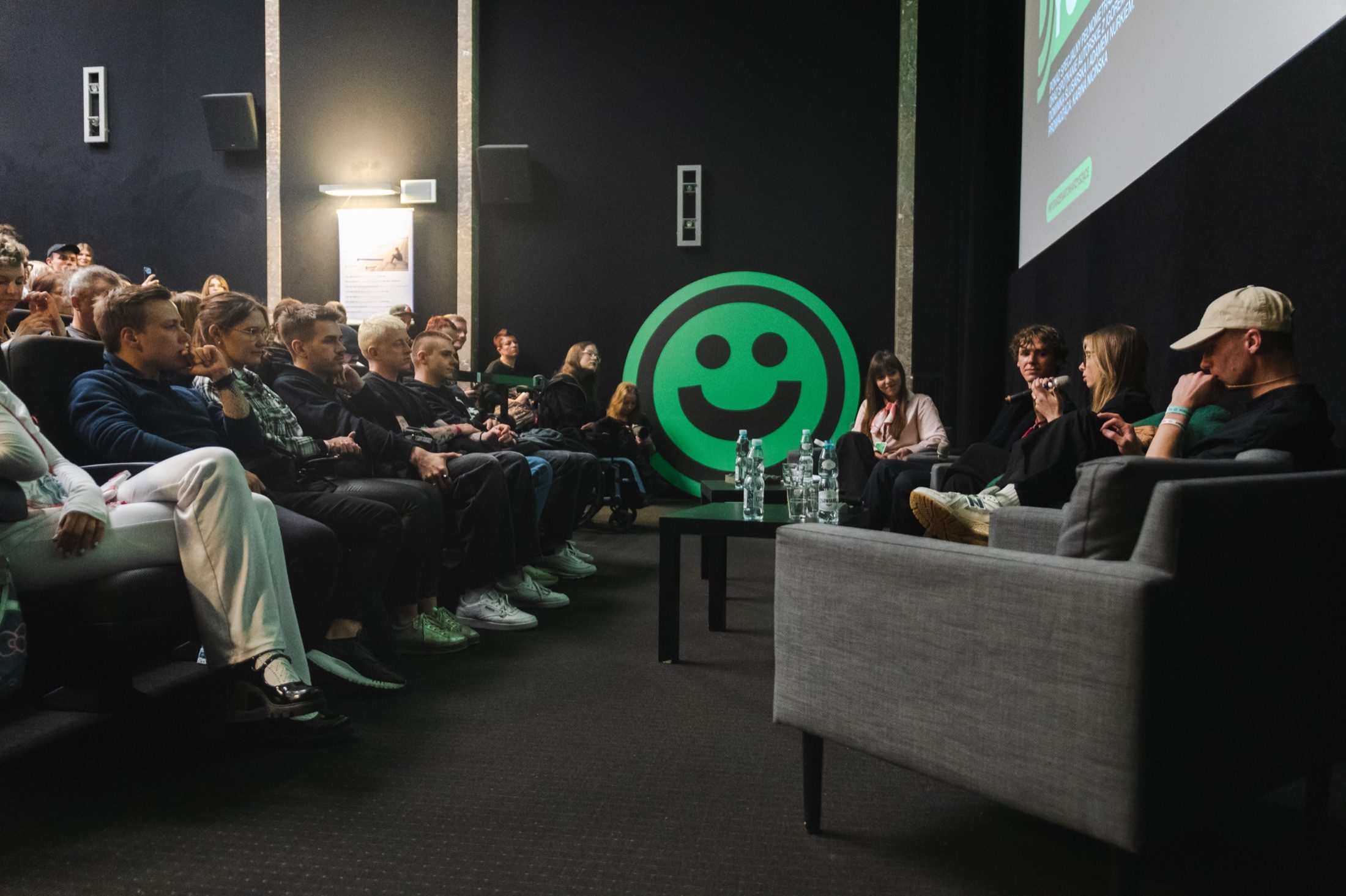 Eine Weitwinkelaufnahme eines Festival-Panels auf der Bühne, das Publikum links und ein großes Festivallogo auf der Leinwand.