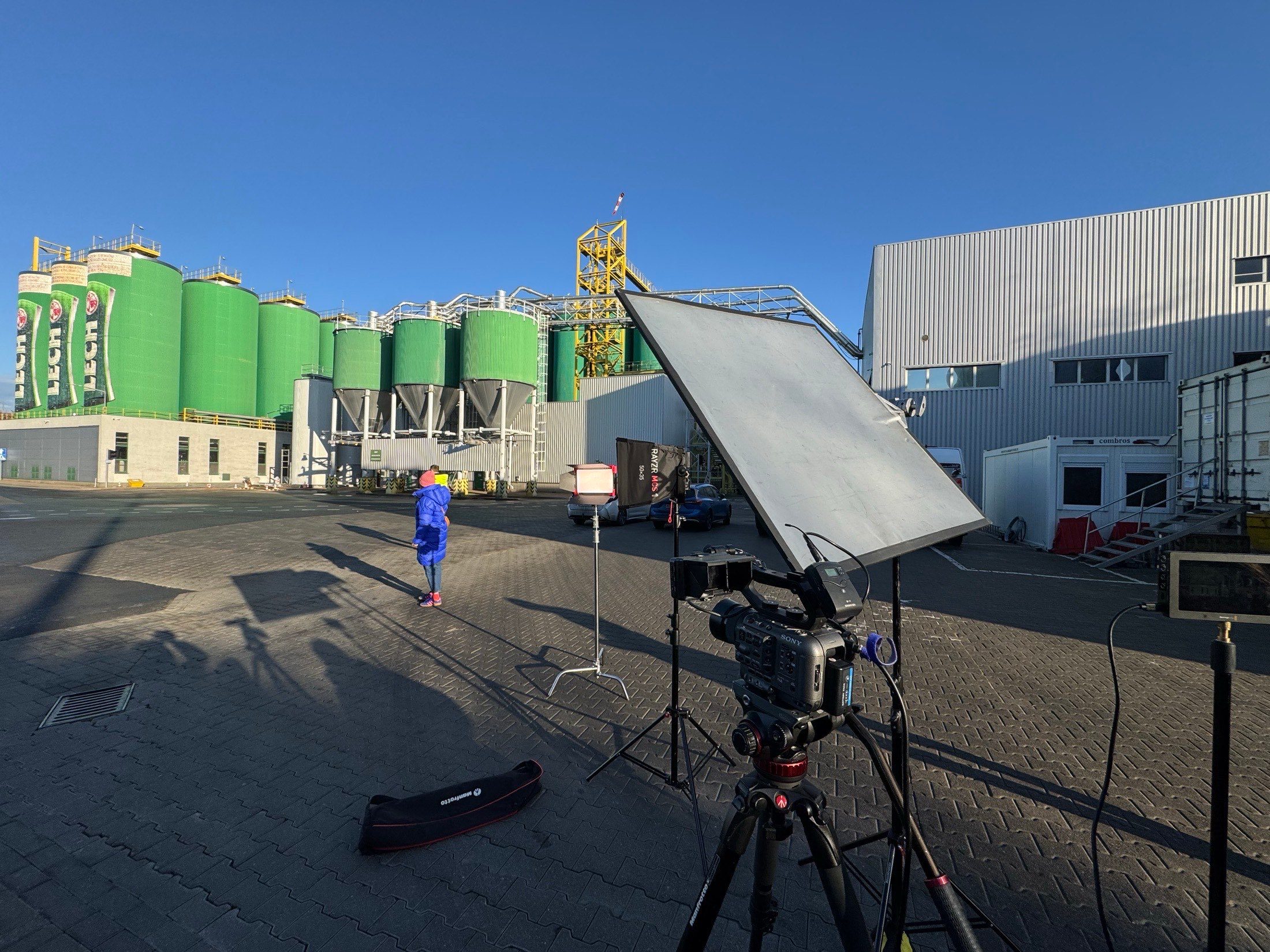 A corporate shoot at an industrial plant: a camera on a tripod and a reflector set up in front of green storage silos under a clear sky.